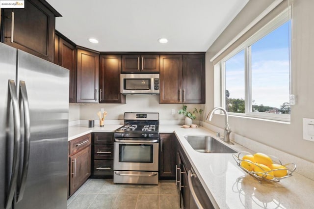 kitchen with appliances with stainless steel finishes, sink, light tile patterned floors, dark brown cabinetry, and light stone countertops