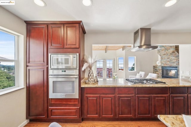 kitchen featuring stainless steel appliances, light stone counters, island range hood, a fireplace, and light wood-type flooring