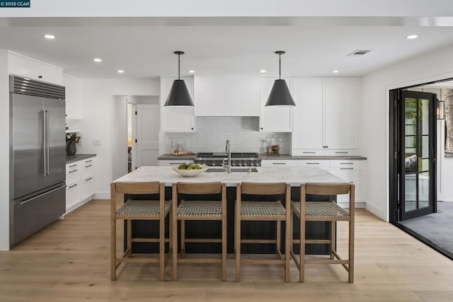 kitchen featuring pendant lighting, stainless steel built in refrigerator, white cabinets, a center island with sink, and light wood-type flooring