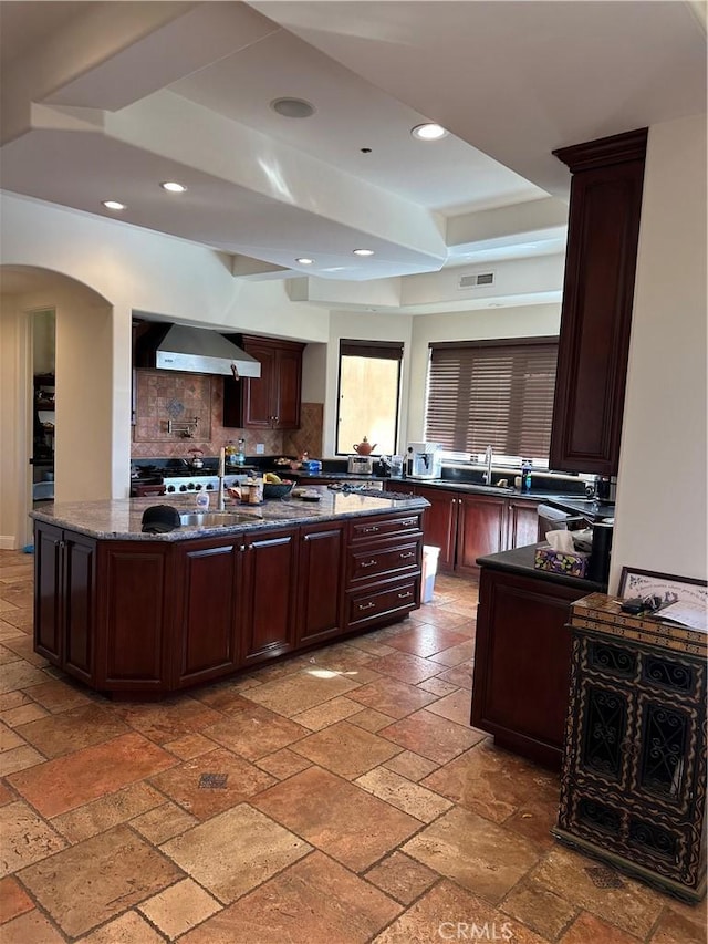 kitchen featuring stone counters, a kitchen island with sink, backsplash, dark brown cabinets, and wall chimney exhaust hood