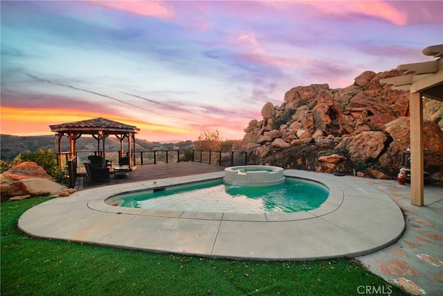 pool at dusk with a wooden deck, a gazebo, and an in ground hot tub