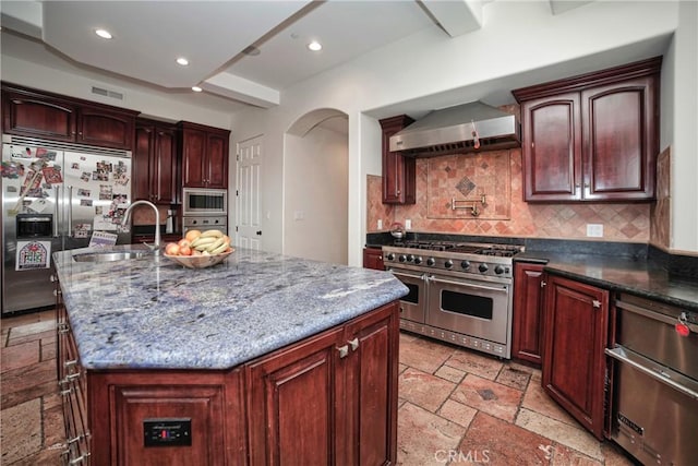 kitchen featuring an island with sink, wall chimney exhaust hood, built in appliances, and sink