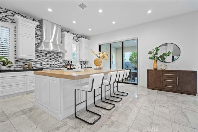 kitchen with white cabinetry, an island with sink, a breakfast bar area, wooden counters, and wall chimney exhaust hood