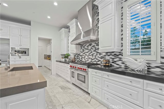 kitchen with wall chimney range hood, sink, premium appliances, white cabinets, and decorative backsplash