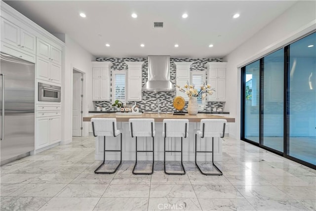 kitchen featuring white cabinets, built in refrigerator, butcher block countertops, and wall chimney range hood