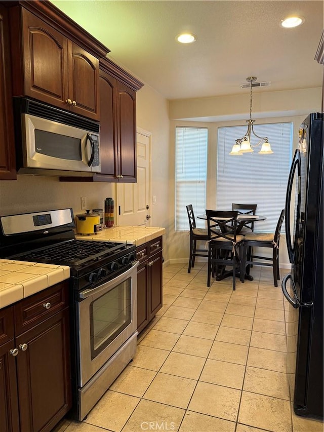 kitchen with light tile patterned floors, dark brown cabinets, stainless steel appliances, decorative light fixtures, and tile countertops