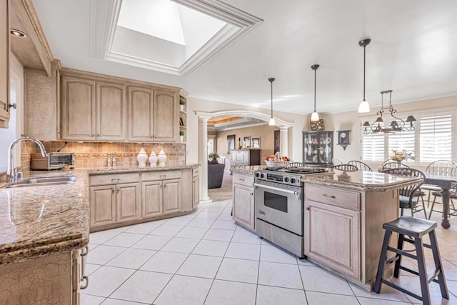 kitchen with light brown cabinetry, sink, high end stainless steel range oven, hanging light fixtures, and a center island