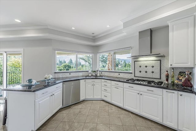 kitchen featuring wall chimney range hood, dark stone counters, white cabinets, and appliances with stainless steel finishes