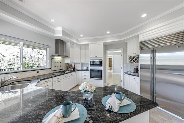 kitchen with wall chimney exhaust hood, sink, white cabinetry, tasteful backsplash, and built in appliances