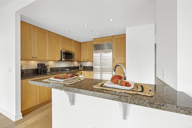 kitchen featuring light wood finished floors, stainless steel appliances, backsplash, dark stone countertops, and a kitchen breakfast bar