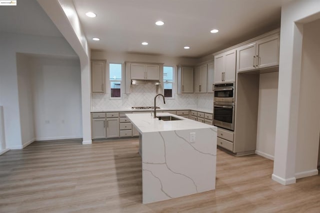 kitchen featuring sink, gray cabinetry, a center island with sink, double oven, and light stone countertops