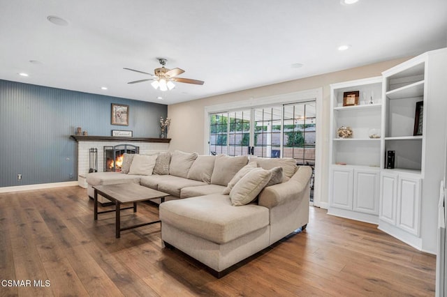 living room with built in shelves, ceiling fan, hardwood / wood-style floors, and a brick fireplace