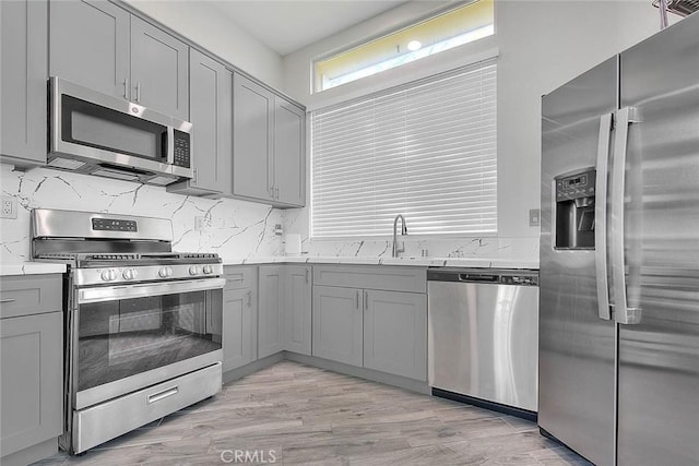 kitchen featuring stainless steel appliances, backsplash, gray cabinetry, a sink, and light wood-type flooring