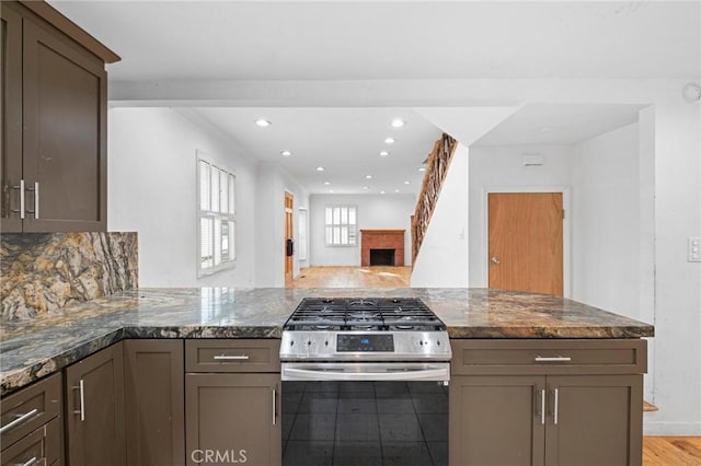 kitchen with tasteful backsplash, dark stone counters, kitchen peninsula, a brick fireplace, and stainless steel range with gas stovetop