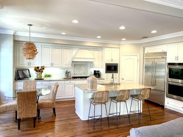 kitchen featuring a kitchen island with sink, built in appliances, tasteful backsplash, decorative light fixtures, and custom exhaust hood