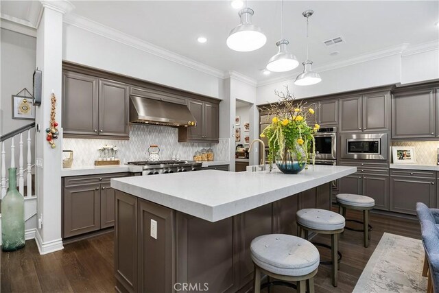 kitchen with an island with sink, pendant lighting, stainless steel appliances, decorative backsplash, and exhaust hood