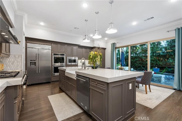 kitchen with sink, built in appliances, decorative light fixtures, an island with sink, and wall chimney range hood