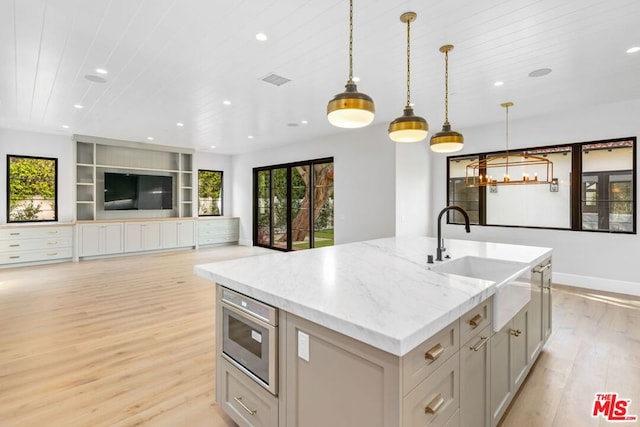 kitchen with sink, hanging light fixtures, light stone counters, a center island with sink, and light wood-type flooring