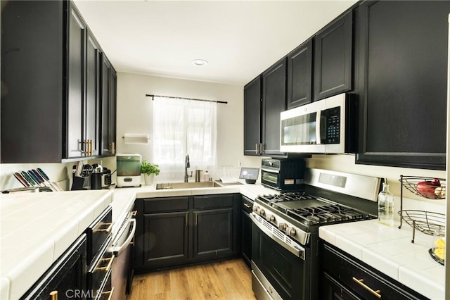 kitchen with appliances with stainless steel finishes, sink, tile countertops, and light hardwood / wood-style flooring