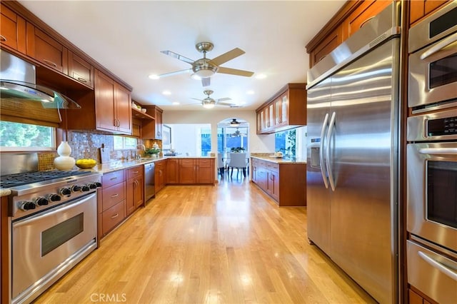 kitchen featuring light wood-type flooring, backsplash, appliances with stainless steel finishes, ceiling fan, and wall chimney range hood