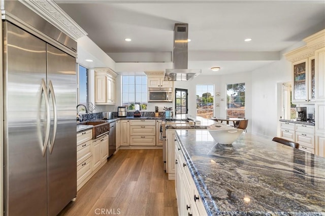 kitchen featuring sink, stainless steel appliances, island range hood, dark stone counters, and light wood-type flooring