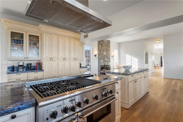 kitchen with stainless steel stove, dark stone countertops, cream cabinets, a fireplace, and wall chimney exhaust hood