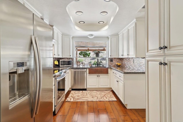 kitchen with a sink, appliances with stainless steel finishes, decorative backsplash, light wood finished floors, and a raised ceiling