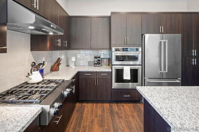 kitchen with appliances with stainless steel finishes, tasteful backsplash, dark brown cabinets, light stone counters, and dark wood-type flooring