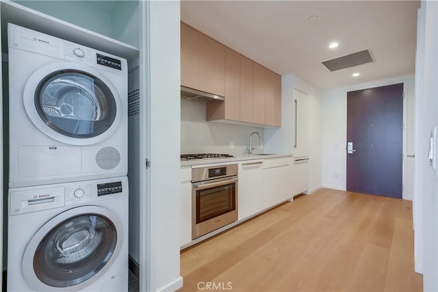 clothes washing area with light wood finished floors, laundry area, recessed lighting, stacked washer / drying machine, and a sink