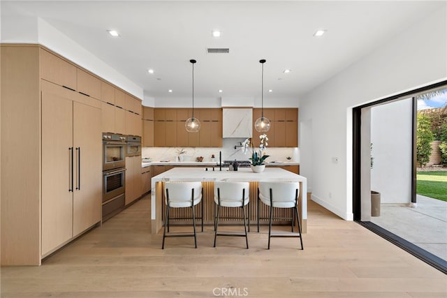 kitchen featuring decorative backsplash, hanging light fixtures, a center island with sink, stainless steel double oven, and light hardwood / wood-style flooring