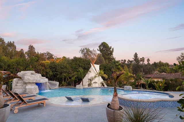 pool at dusk with pool water feature, an in ground hot tub, and a patio