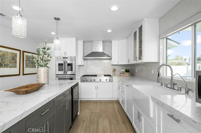 kitchen with appliances with stainless steel finishes, pendant lighting, white cabinetry, light stone counters, and wall chimney range hood