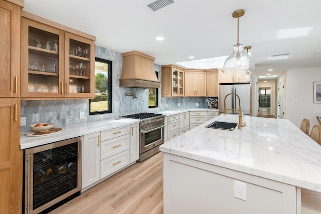 kitchen with appliances with stainless steel finishes, a kitchen island with sink, white cabinets, custom exhaust hood, and beverage cooler