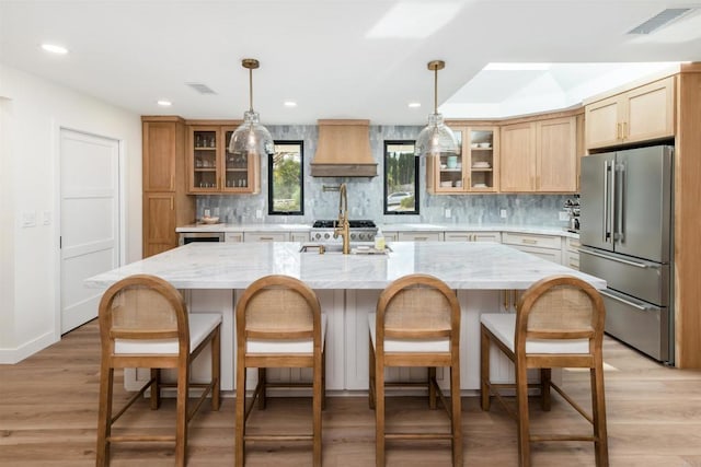 kitchen featuring high end fridge, wall chimney range hood, a kitchen island with sink, and light stone countertops