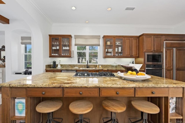 kitchen with sink, built in appliances, light stone counters, ornamental molding, and a kitchen island