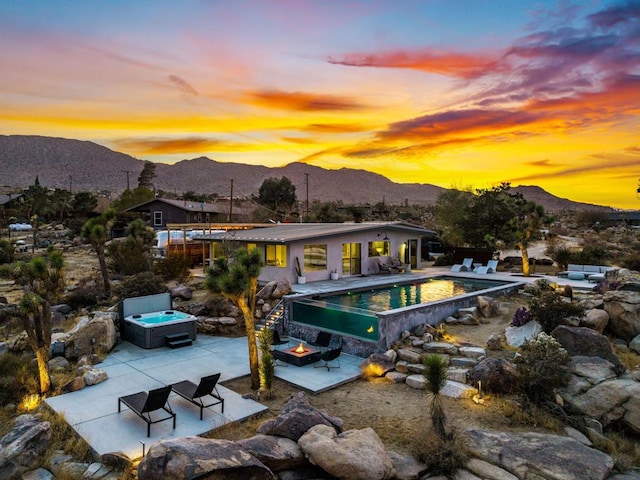 back house at dusk with a mountain view, a pool with hot tub, an outdoor fire pit, and a patio area