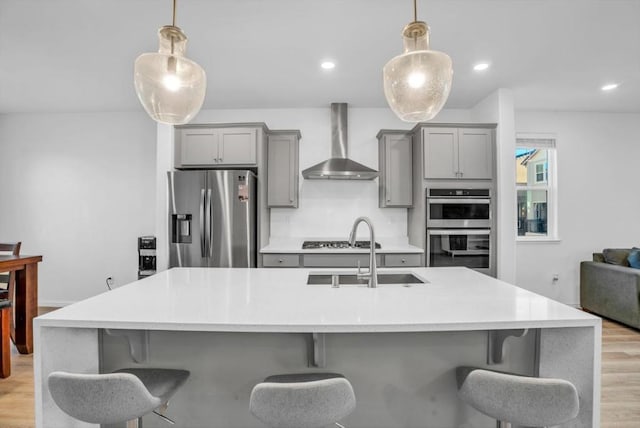 kitchen featuring stainless steel appliances, hanging light fixtures, wall chimney range hood, and a center island with sink