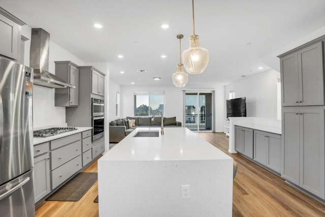 kitchen featuring wall chimney exhaust hood, sink, decorative light fixtures, appliances with stainless steel finishes, and a kitchen island with sink