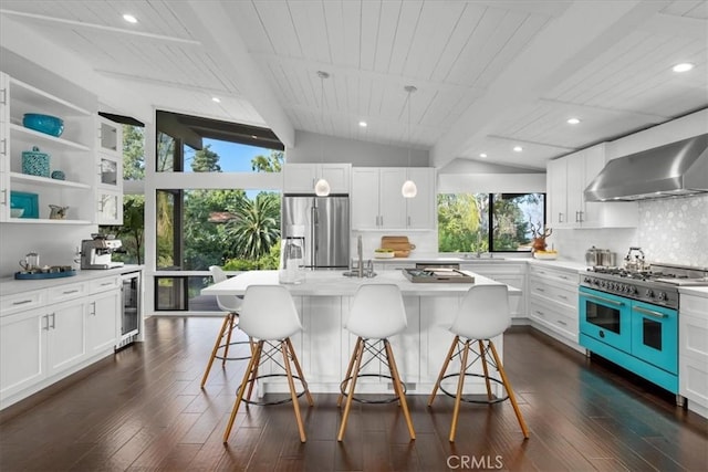 kitchen featuring an island with sink, white cabinets, range with two ovens, stainless steel refrigerator with ice dispenser, and wall chimney exhaust hood