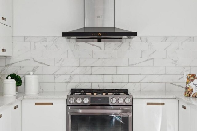 kitchen with stainless steel range with gas stovetop, white cabinets, wall chimney range hood, light stone countertops, and backsplash