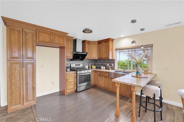 kitchen featuring wall chimney range hood, sink, a kitchen breakfast bar, stainless steel gas range oven, and decorative light fixtures