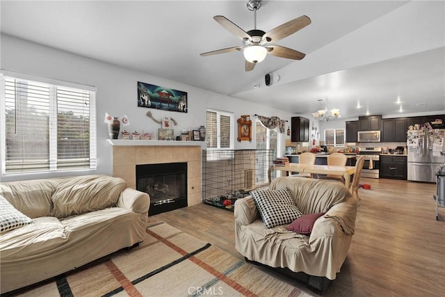 living room featuring lofted ceiling, ceiling fan with notable chandelier, a tile fireplace, and light wood-type flooring