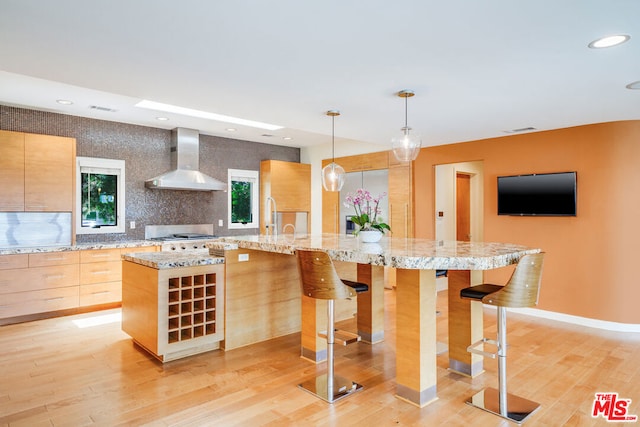 kitchen with tasteful backsplash, hanging light fixtures, a kitchen island with sink, light stone countertops, and wall chimney range hood