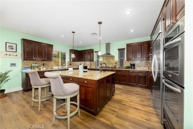 kitchen with a center island with sink, a kitchen breakfast bar, hanging light fixtures, light wood-type flooring, and wall chimney range hood