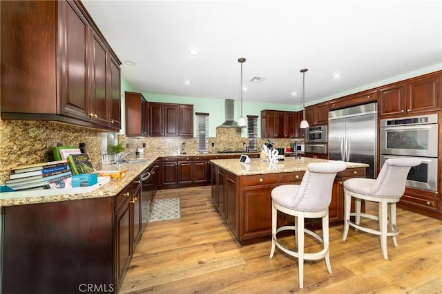 kitchen featuring pendant lighting, appliances with stainless steel finishes, wall chimney range hood, an island with sink, and a kitchen breakfast bar