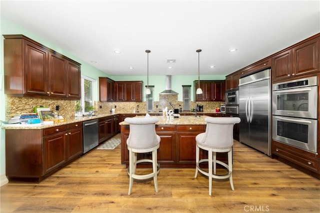 kitchen featuring decorative light fixtures, a center island with sink, appliances with stainless steel finishes, wall chimney range hood, and a kitchen bar