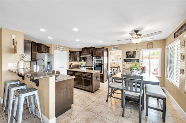 kitchen featuring light tile patterned floors, appliances with stainless steel finishes, dark brown cabinetry, light stone counters, and kitchen peninsula