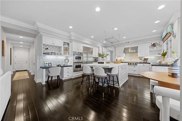 interior space with a breakfast bar, built in appliances, tasteful backsplash, white cabinets, and wall chimney exhaust hood