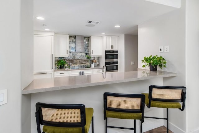 kitchen featuring double oven, a breakfast bar area, white cabinets, backsplash, and wall chimney exhaust hood