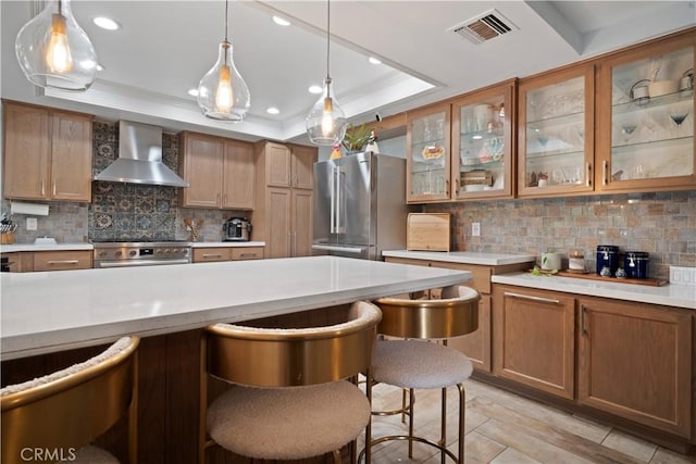 kitchen with appliances with stainless steel finishes, decorative light fixtures, a kitchen breakfast bar, a raised ceiling, and wall chimney exhaust hood